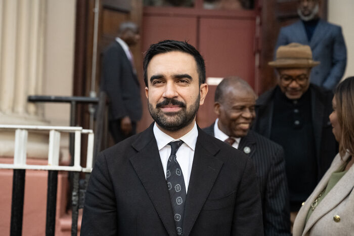 Zohran Mamdani wearing a suit, standing outdoors with people in background at a public event.