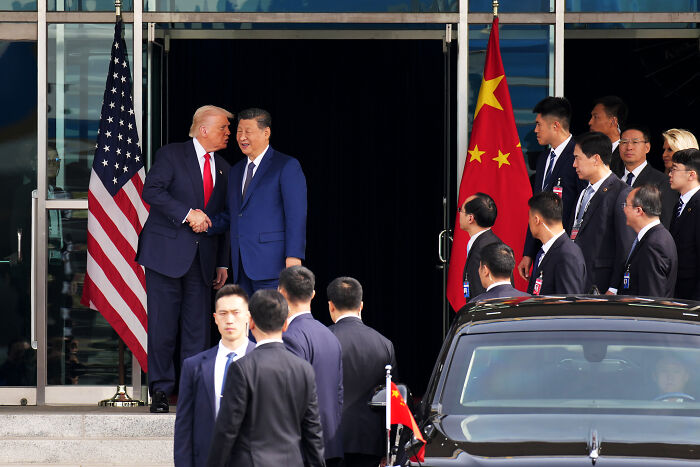 Donald Trump shaking hands with Xi Jinping outside a building, with officials and flags of the US and China present.