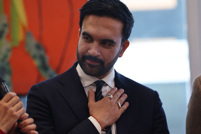 Zohran Mamdani wearing a dark suit, touching his chest thoughtfully during a discussion about democratic socialism.