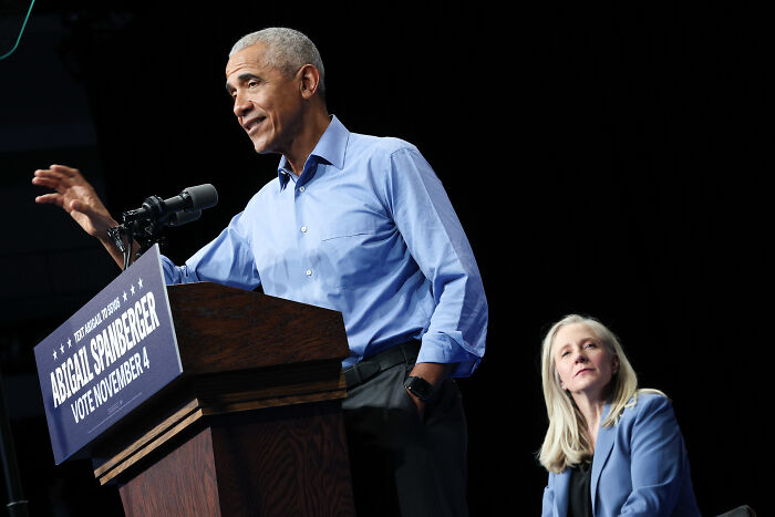 Barack Obama speaking at a podium, gesturing with hand, while a woman in blue listens attentively during an event.