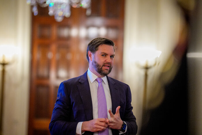 Man in suit and purple tie speaking indoors with wooden doors behind, related to Trump comments on JD Vance social media reaction
