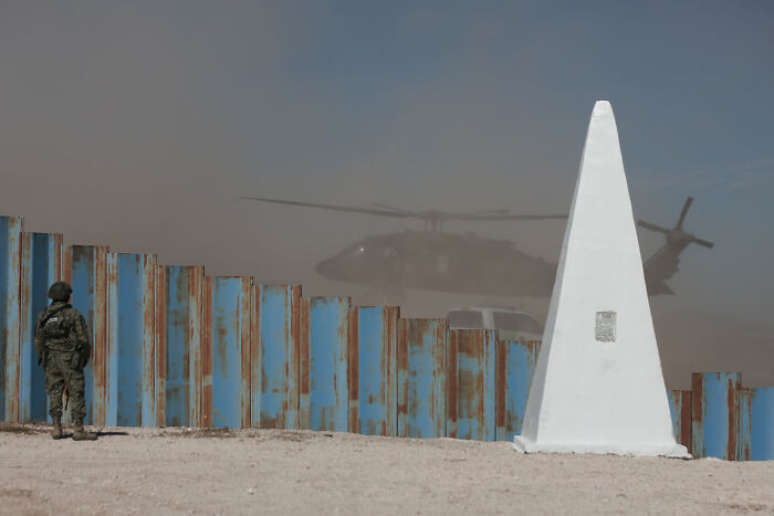 Soldier standing by a rusted border fence with a helicopter landing nearby, illustrating migration control and surveillance states.