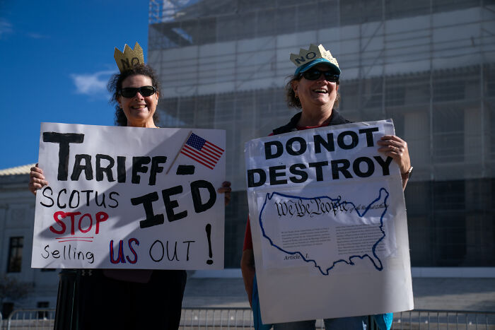 Two women wearing crowns hold protest signs expressing concern over tariffs and constitutional rights in Washington D.C.