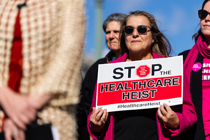 Protester holding sign at rally, highlighting healthcare issues amid MAGA son and SNAP Thanksgiving debate.