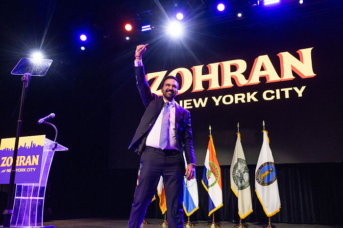 Zohran Mamdani at NYC mayoral victory event raising fist on stage with flags and campaign signage behind him.