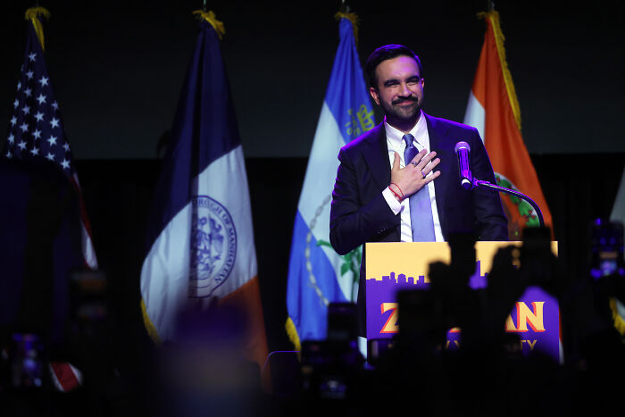 Zohran Mamdani at a podium with flags behind him, addressing audience after winning NYC mayoral race.