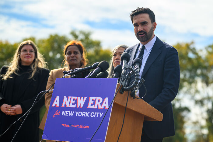 Zohran Mamdani speaking at a podium with a New York City sign, facing media and supporters outdoors.