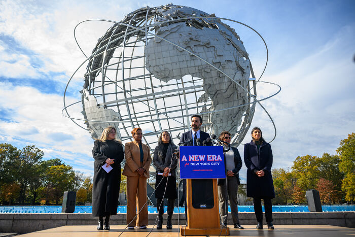Zohran Mamdani speaking at a podium with a globe sculpture behind during NYC mayoral race event.