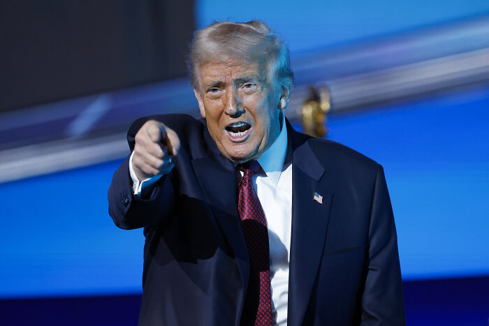 Donald Trump in a dark suit and red tie pointing forward passionately during a public speaking event.