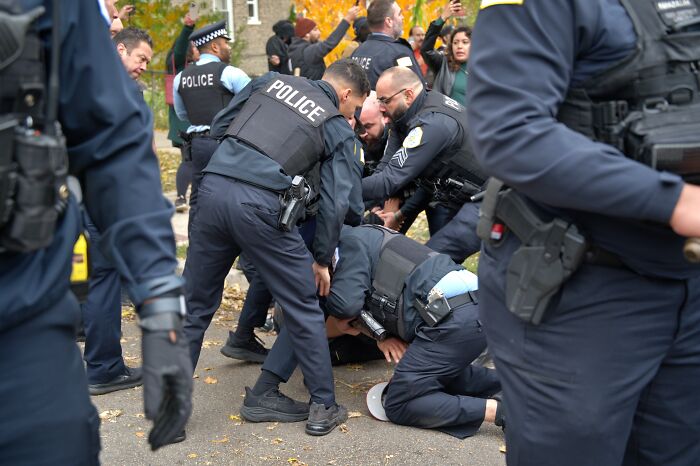 Several police officers restraining a person on the ground during a public protest or confrontation scene outdoors.