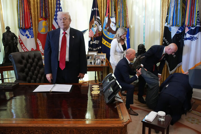 Donald Trump standing with a serious expression while a man faints and is helped by others behind him in the Oval Office.