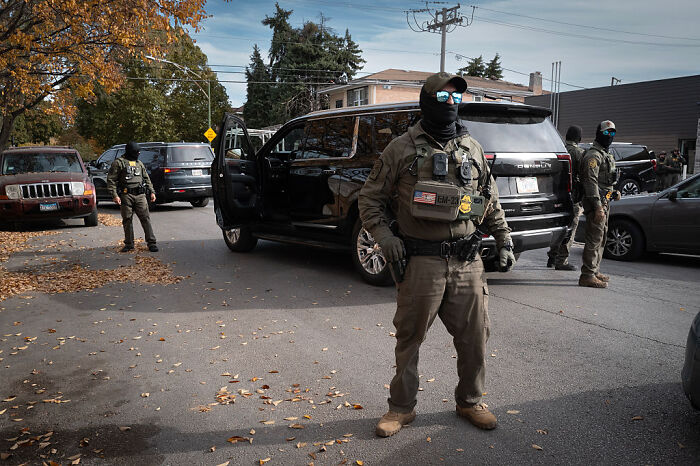 Federal agent wearing tactical gear standing near black SUV on a street during operation in Chicago neighborhood.