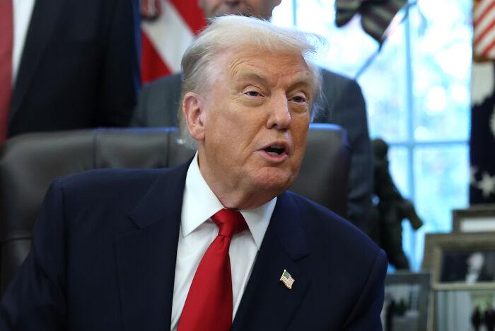 Former President Trump in the Oval Office, speaking during a meeting while wearing a dark suit and red tie.