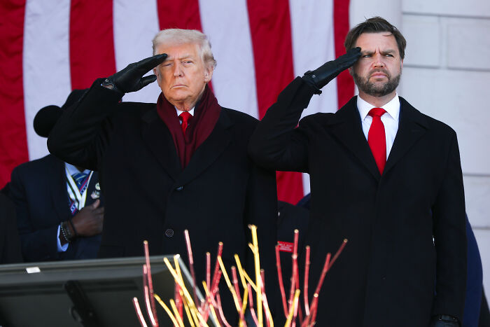 Donald Trump and another man saluting during a Veterans Day event with American flag backdrop.