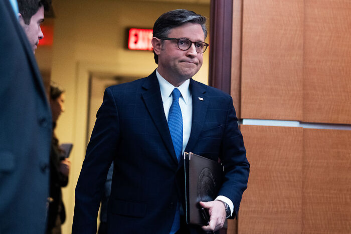 Man in a suit and glasses walking indoors holding a folder, symbolizing political tension and warnings of political violence.