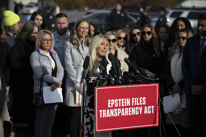 Woman speaking at podium with Epstein Files Transparency Act sign, surrounded by supporters during outdoor press event.