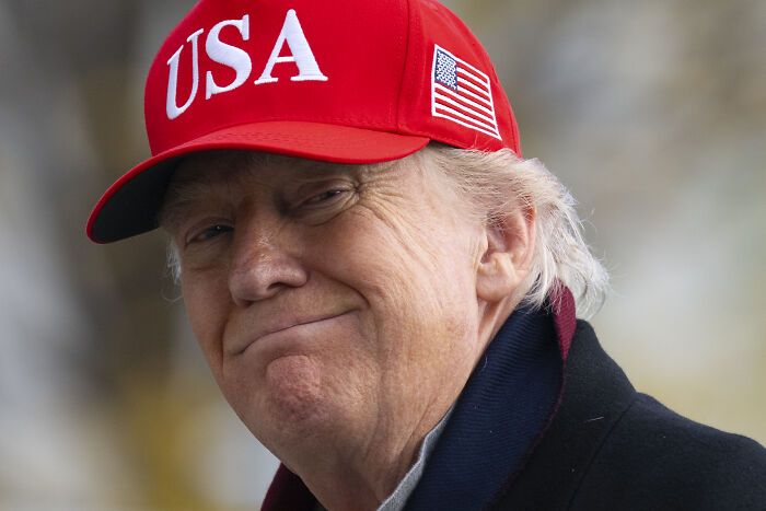 Donald Trump wearing a red USA cap, smiling with a subtle smirk, linked to personality cult and Republican Party rename.