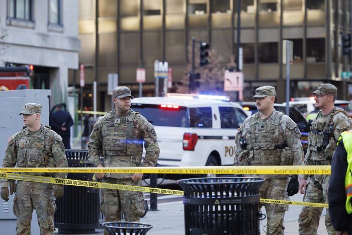 National Guard members stand behind police tape at a D.C. shooting scene with police vehicles in the background.