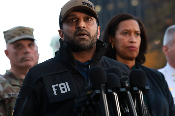 Kash Patel speaking at press conference, wearing FBI jacket and cap, addressing National Guard gunman situation with officials behind him.