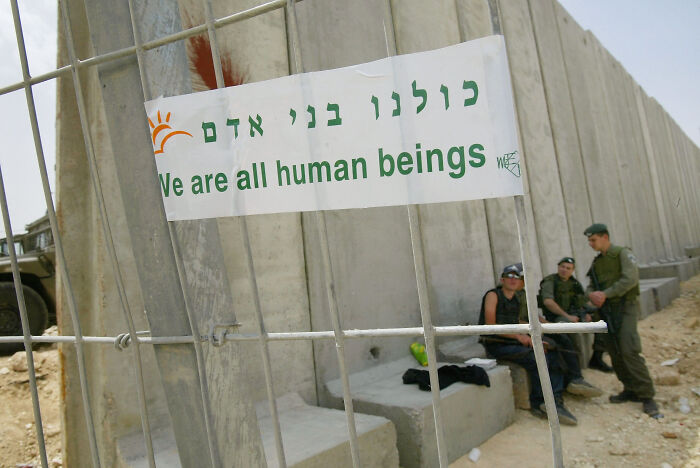 Banner on metal fence reads We are all human beings near concrete barrier with soldiers, illustrating migration control and surveillance states.