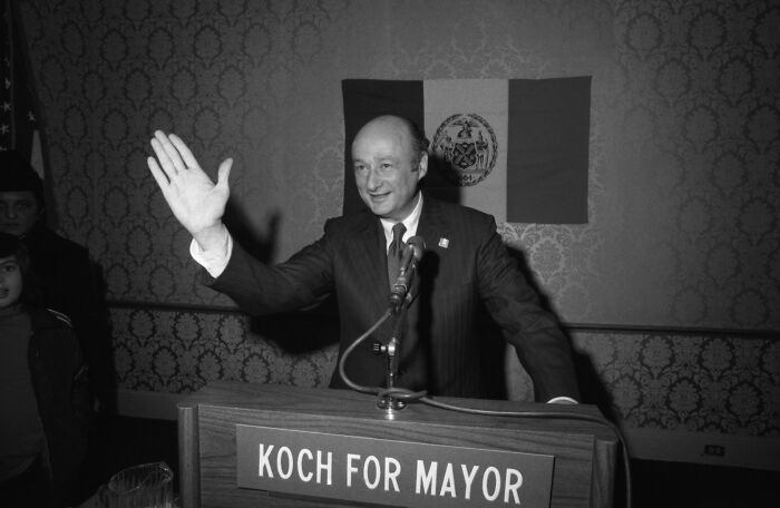 Black and white photo of a politician at a podium with Koch for Mayor sign, related to Trump piggy insult New York skyscraper battle.