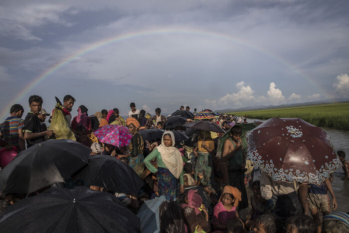 Crowd of migrants with umbrellas under a rainbow, illustrating migration control in surveillance states and data clouds.