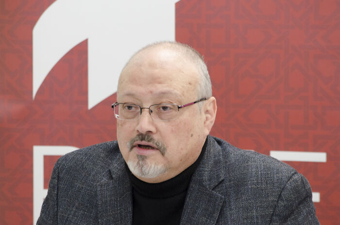 Jamal Khashoggi speaking at an event, wearing glasses and a dark blazer with a patterned red background.