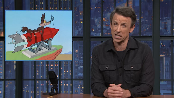 Late-night host Seth Meyers delivering a monologue with a cartoon of Wile E. Coyote on a rocket in the background.