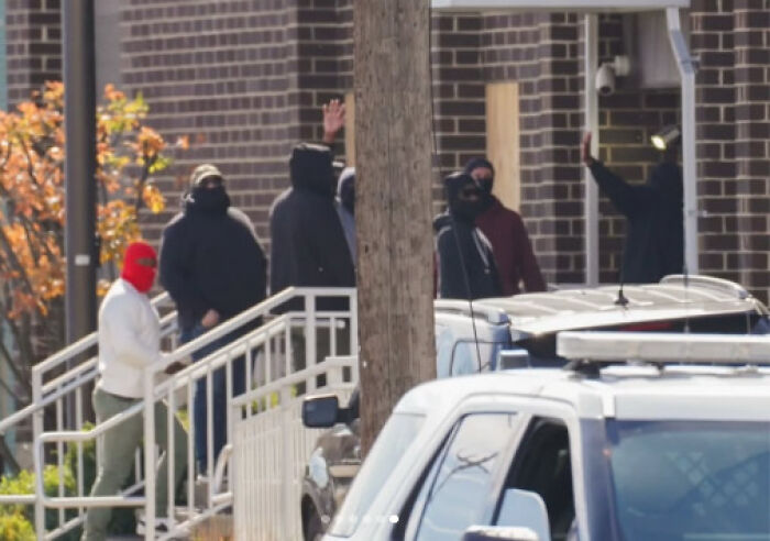 Man in tears runs to follow disappeared wife at ICE facility, captured outside a brick building with masked individuals nearby.