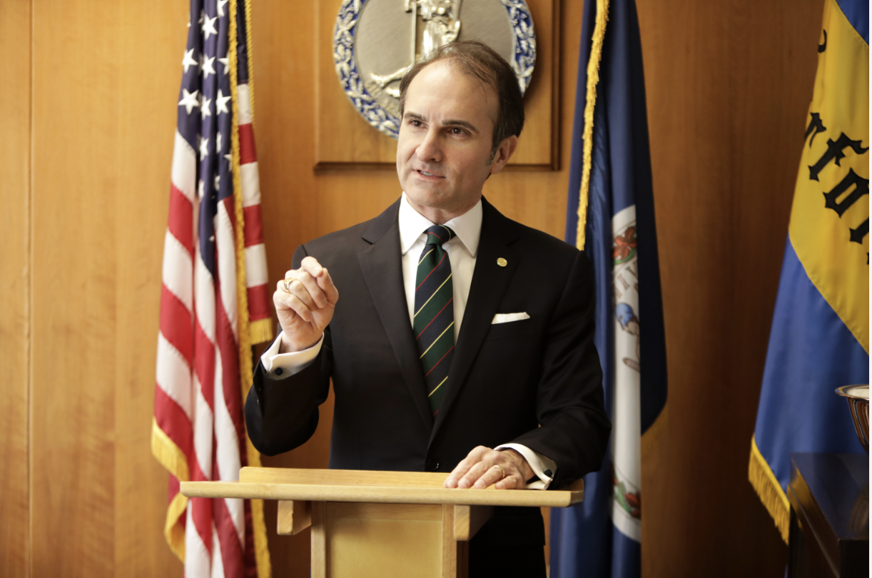 Man in suit speaking at a podium with American and Virginia state flags in the background about parents jailed and baby stroller fall case