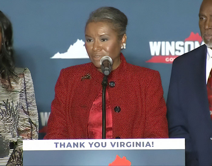 Female speaker at podium with thank you Virginia sign, highlighting Virginia turning blue in a sweeping victory against Trump.