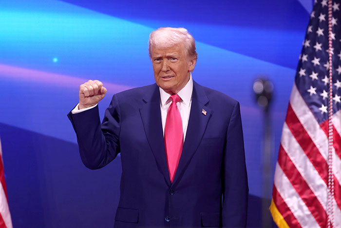 Donald Trump raising fist at a podium with American flags in the background during a political event.