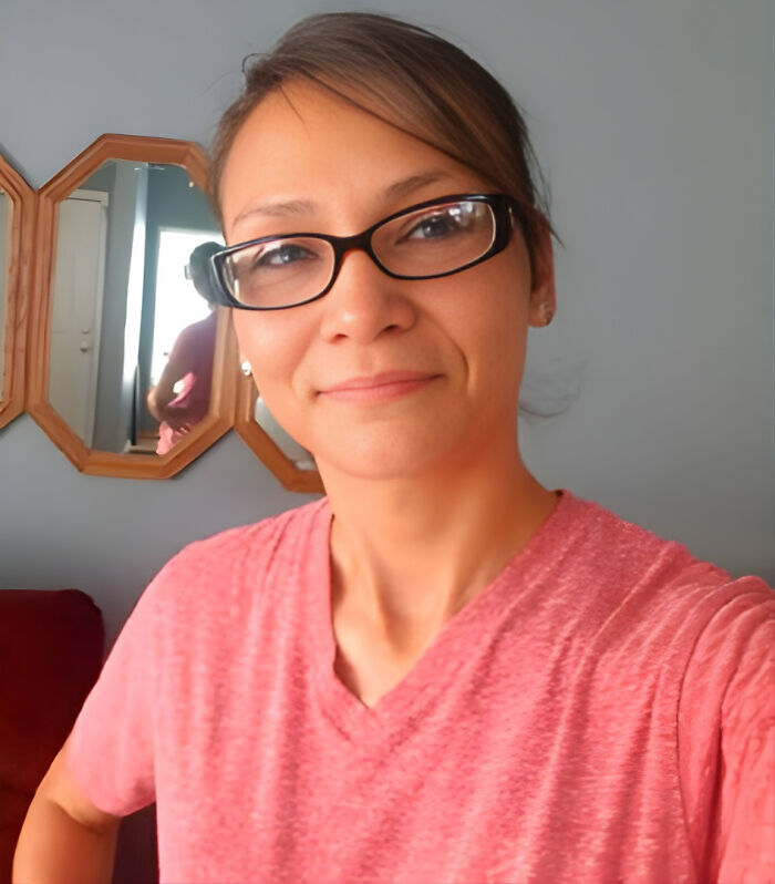 Indigenous woman wearing glasses and a pink shirt, smiling indoors with mirrors on the wall behind her.
