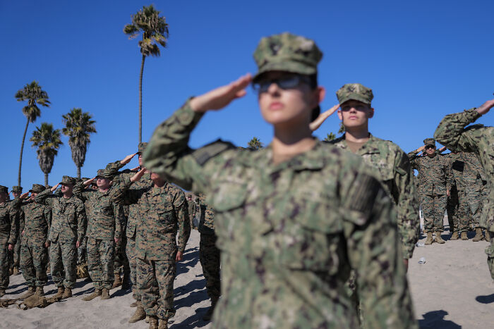 U.S. military personnel in camouflage uniforms salute during an outdoor event under clear blue sky with palm trees nearby.