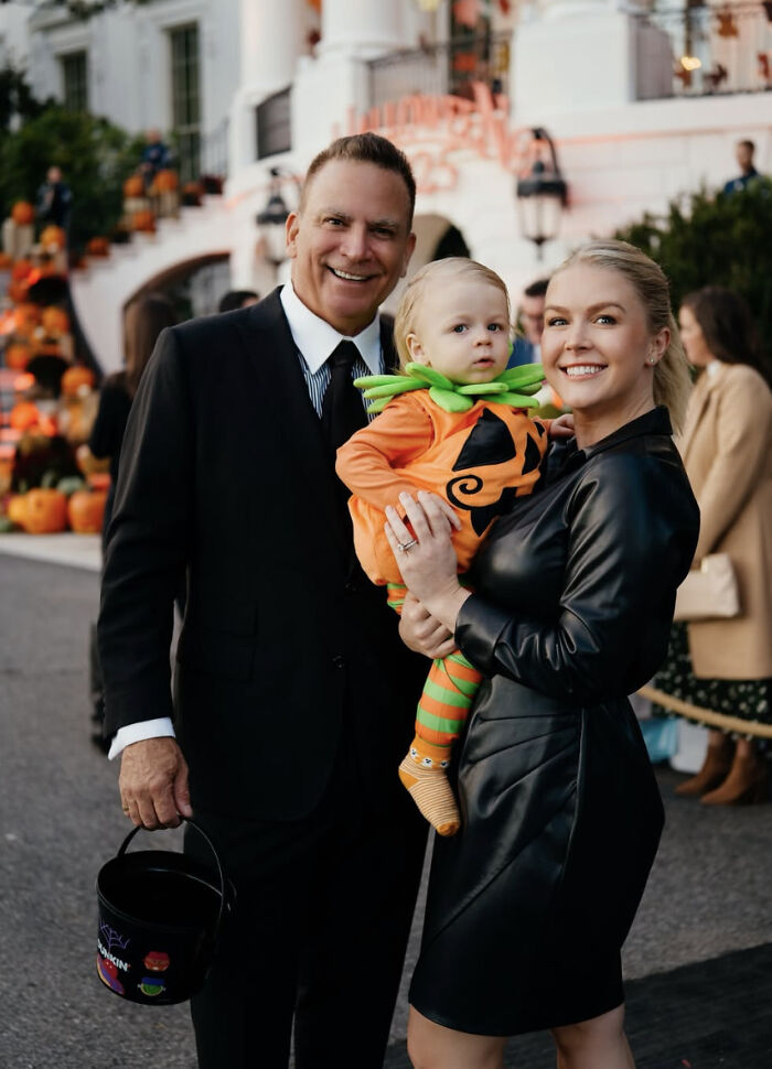 Karoline Leavitt smiling in black dress holding child in pumpkin costume with husband in suit during Halloween event.