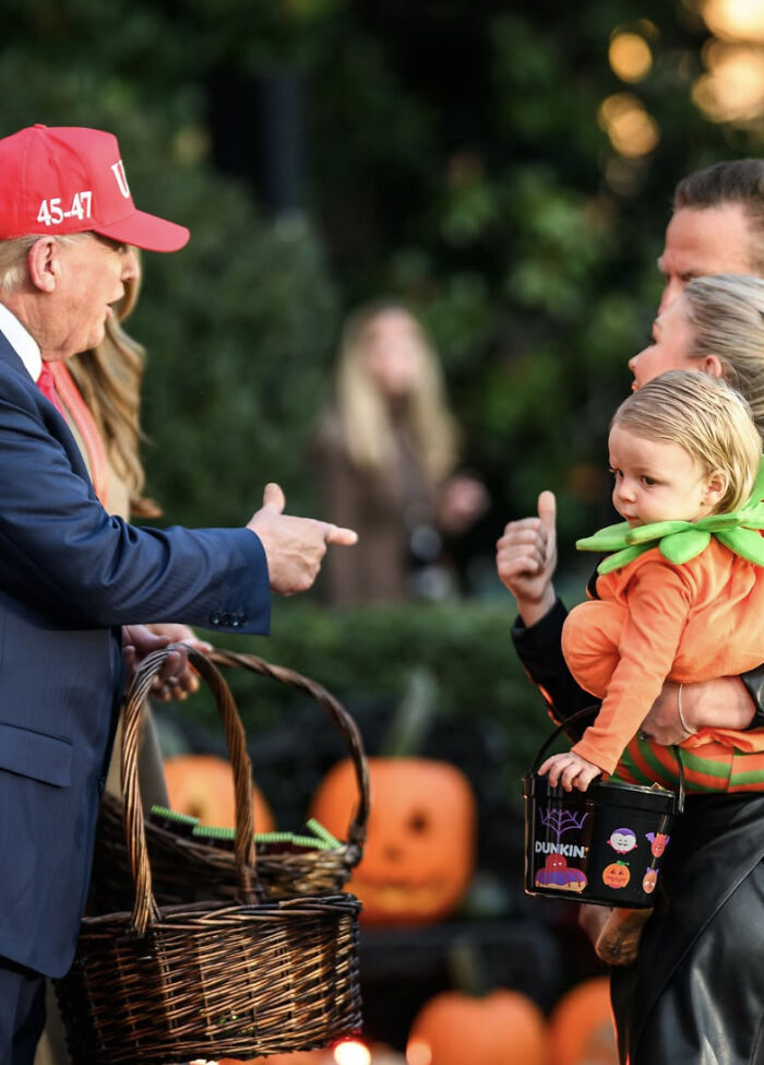 Man in red hat handing out Halloween treats while woman holds child in pumpkin costume during outdoor event