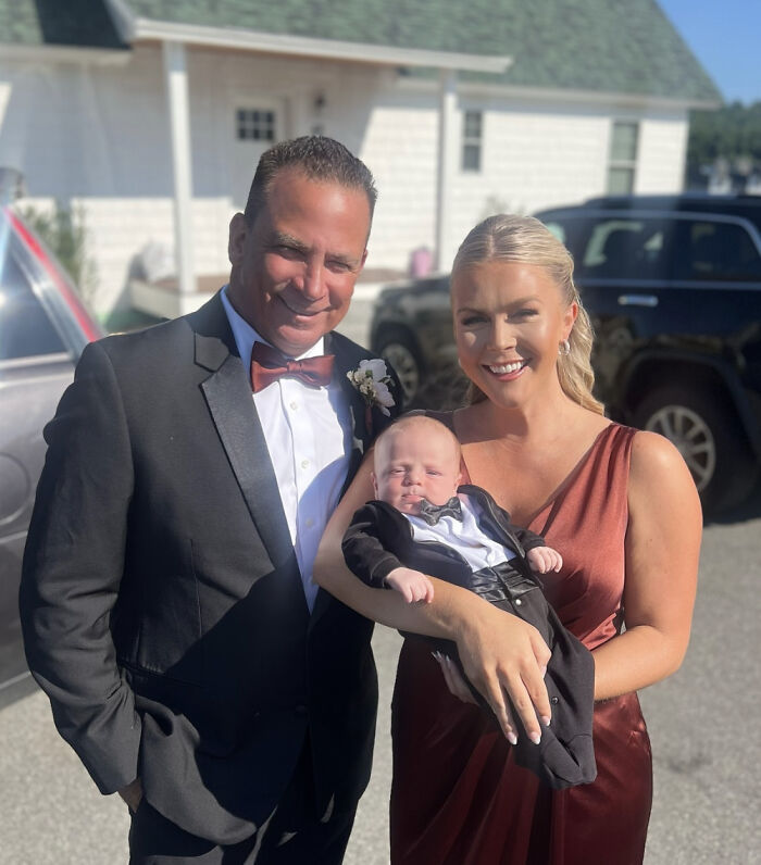 Karoline Leavitt in a red dress holding a baby, standing next to a man in a black tuxedo, smiling outdoors on a sunny day.