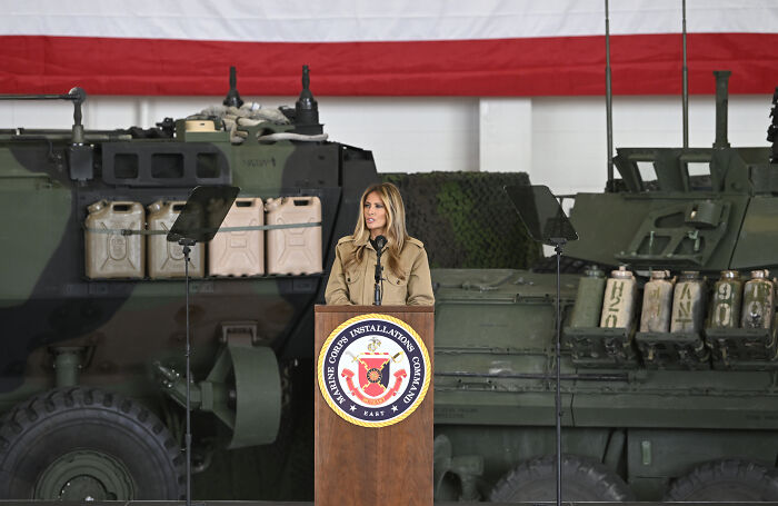 Melania Trump speaking at military podium with armored vehicles in background during troops address event.