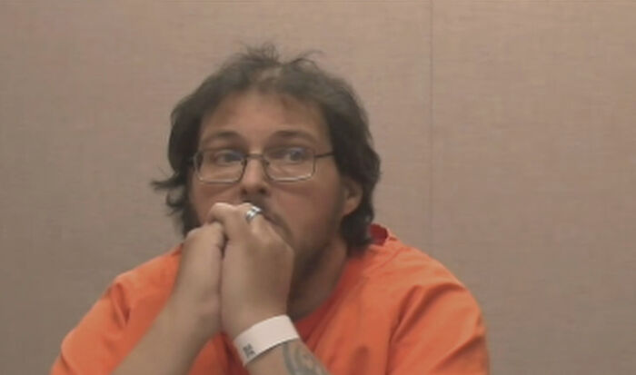 Man in an orange prison jumpsuit with glasses, looking anxious and resting his hands on his mouth in a plain room.