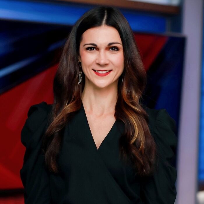 TV sports reporter smiling in studio with long dark hair and black blouse, news set background.