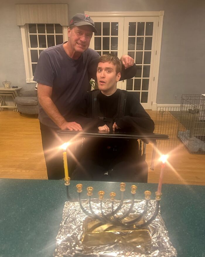 Man standing beside his disabled son in a wheelchair, with a lit menorah in the foreground, discussing Trump&rsquo;s nephew.