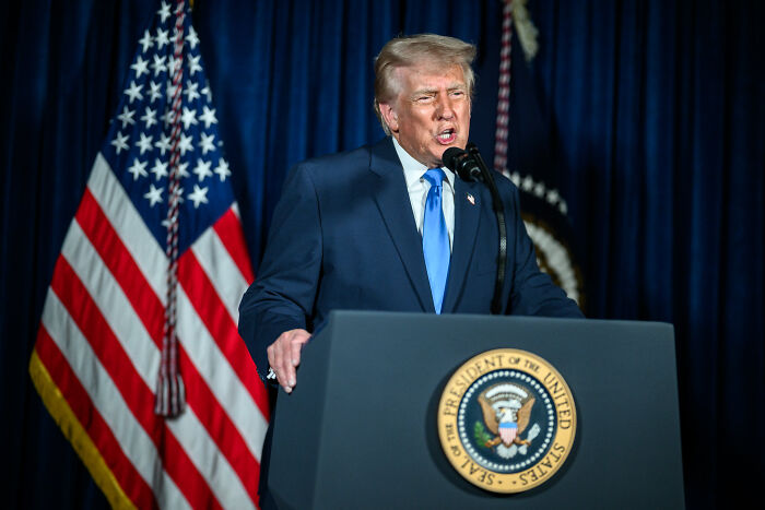 Former President Trump speaking at a podium with the presidential seal, flanked by American flags, amid MAGA base division.
