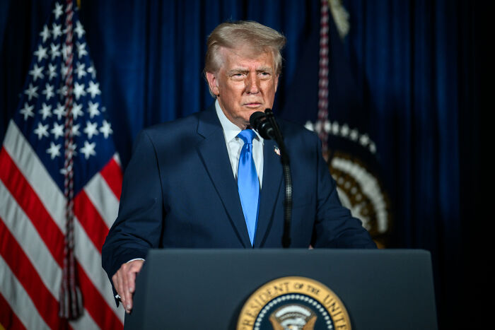 Donald Trump speaking at a podium with the presidential seal, American flag, and presidential emblem in the background.