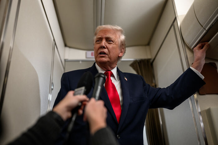 Former President Trump speaking inside an aircraft, surrounded by reporters with microphones during a media interaction.