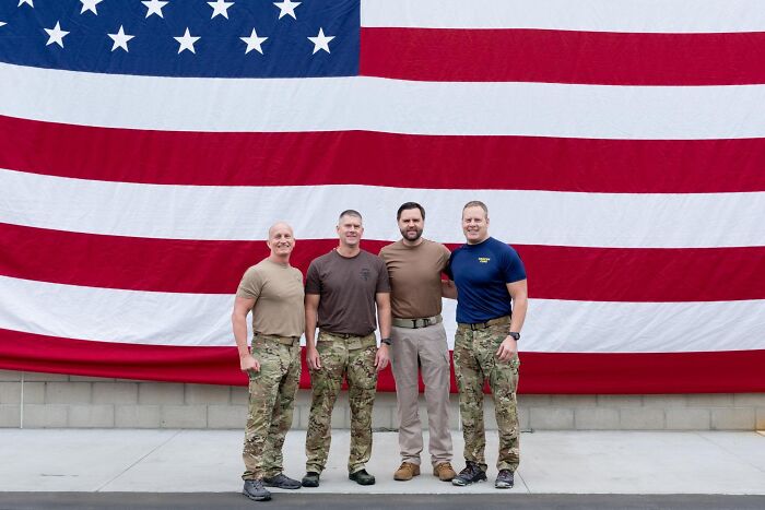 Four men in military and civilian clothing standing in front of a large American flag referencing Navy SEAL cosplay controversy.