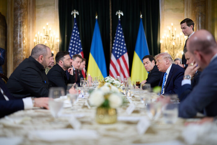 Volodymyr Zelenskyy and Donald Trump seated at a formal meeting table with Ukrainian and American flags in the background.