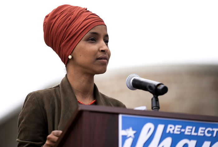 Ilhan Omar speaking at a podium outdoors wearing a red headwrap, addressing issues involving ICE and Somali immigrants.