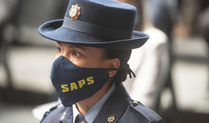 South African police officer wearing SAPS face mask and uniform at a briefing on fatal mass shooting victims including children and teens.