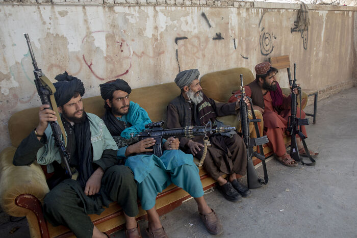 Four armed Taliban fighters sitting on a couch holding rifles against a wall in a desert setting.