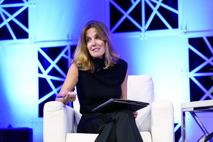 Woman in black sleeveless top sitting in white chair speaking about corporate censorship at media event with blue backdrop.